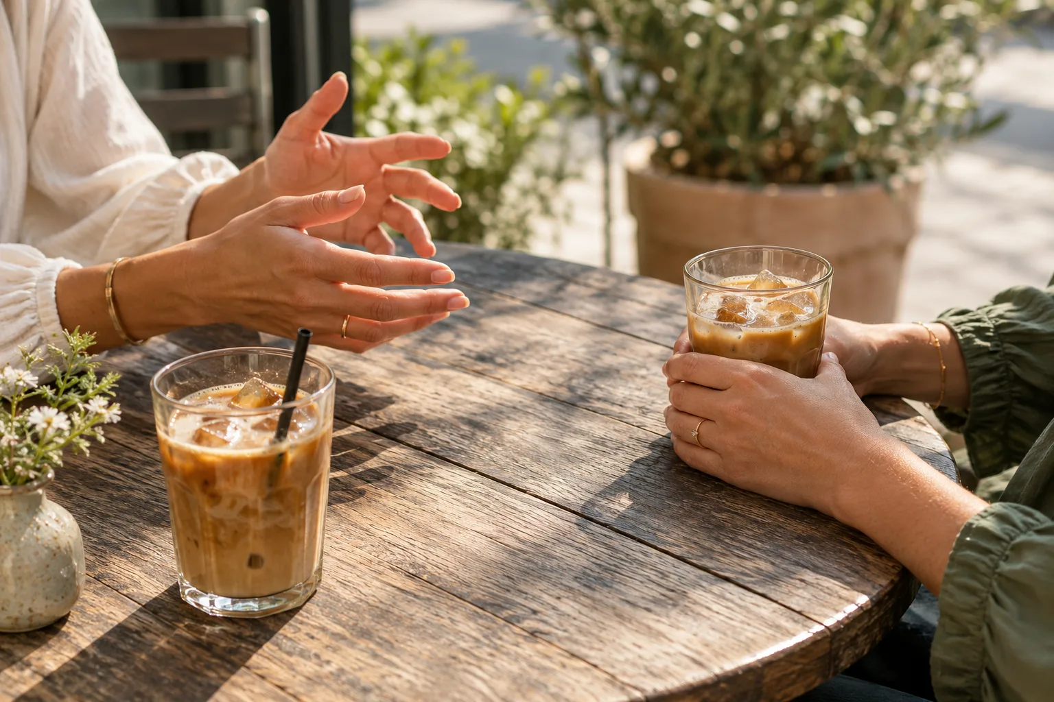 Two people's hands on a cafe table, one gesturing while talking, the other holding an iced coffee
