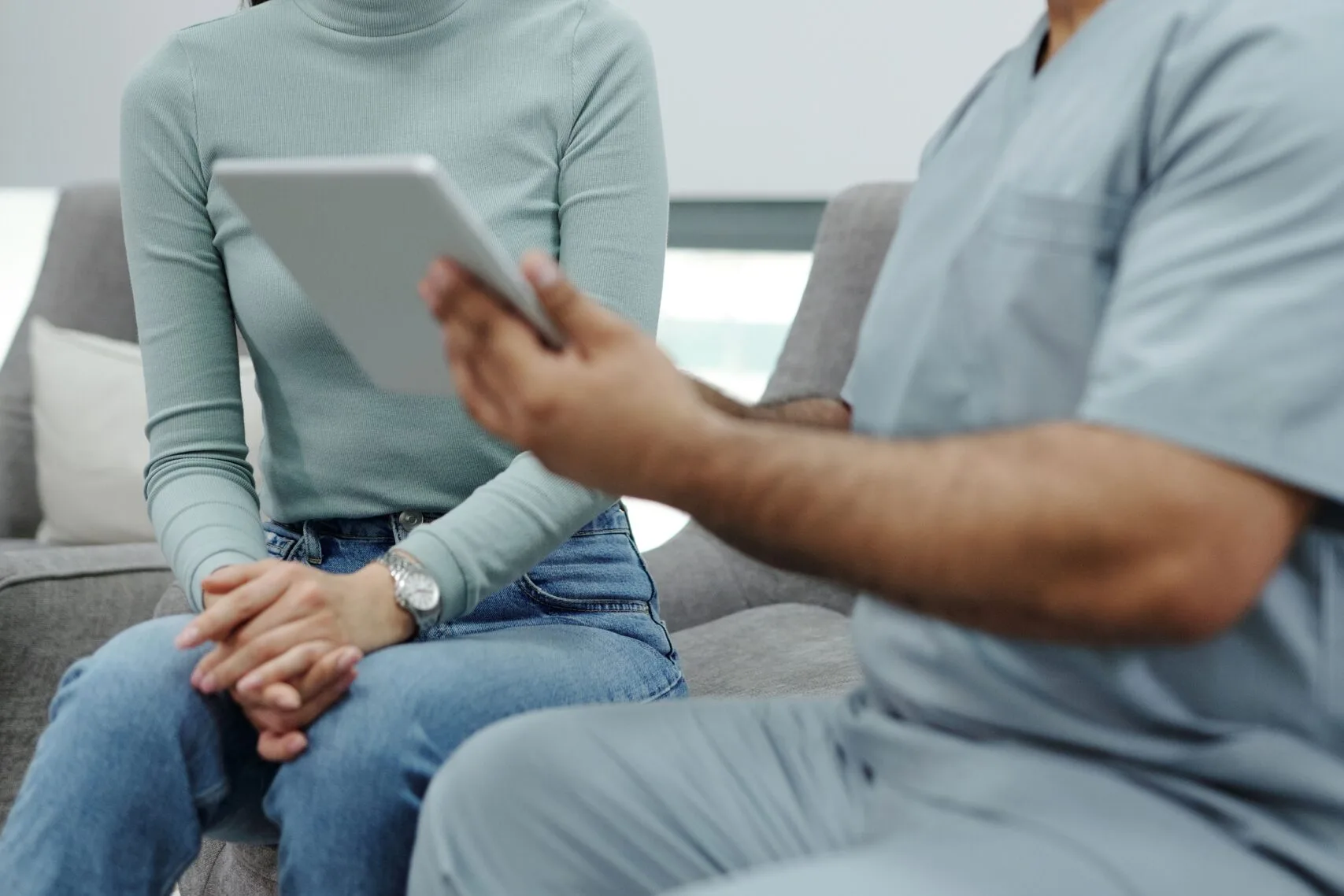 A seated patient with hands clasped while a clinician in scrubs holds a tablet
