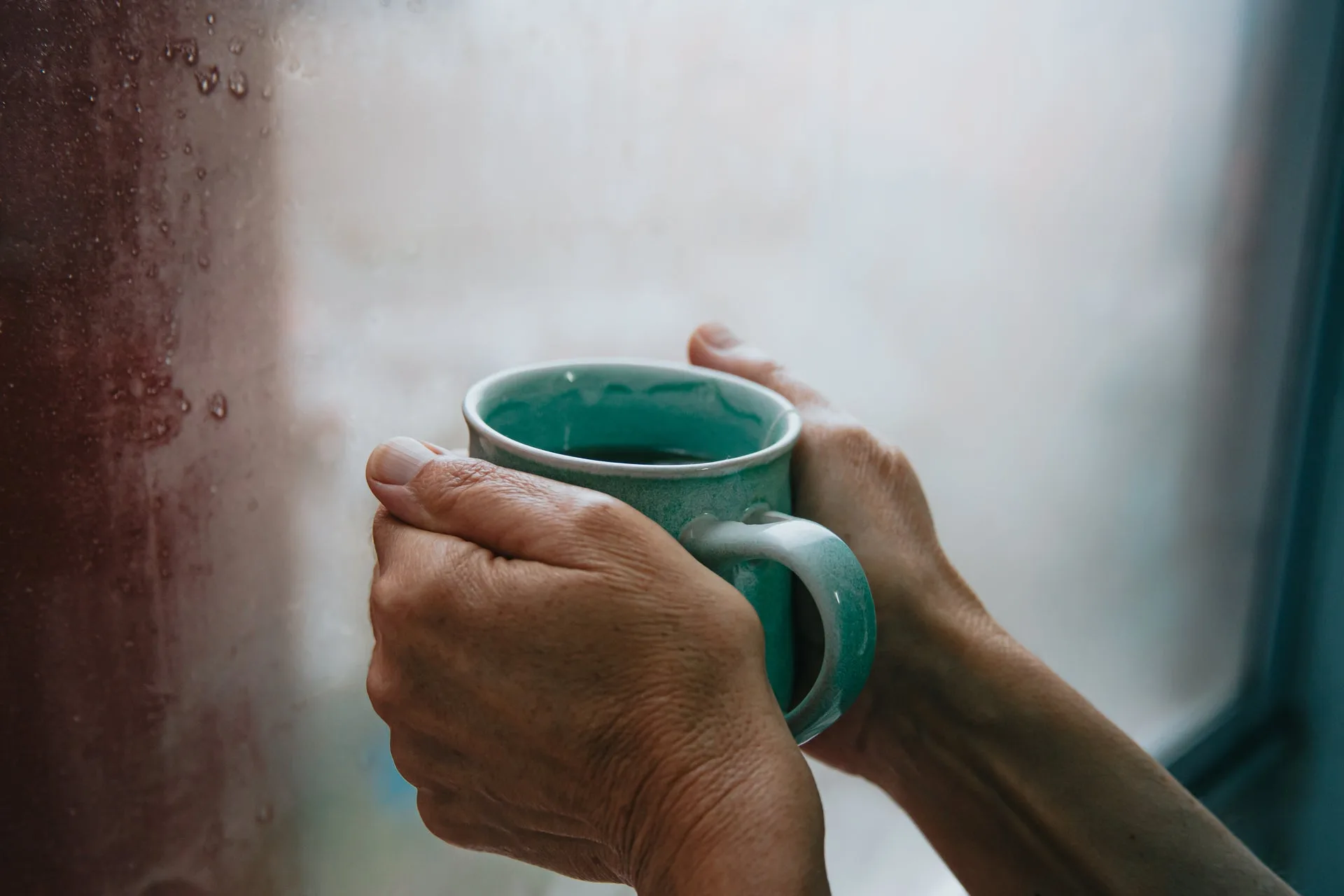 Two hands holding a green ceramic mug beside a rain-streaked window