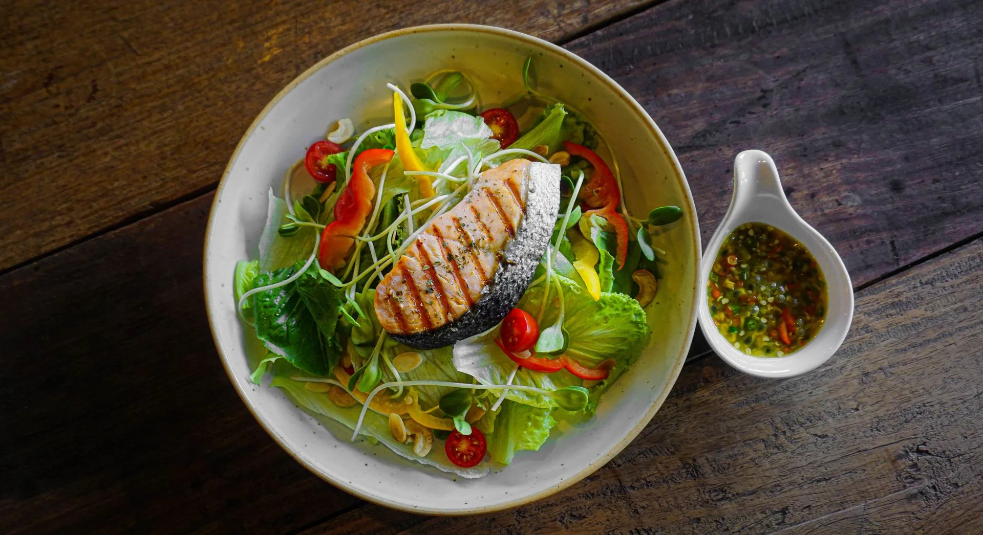 A bowl of salad with a grilled salmon steak and a small dish of dressing on a wooden table, seen from above