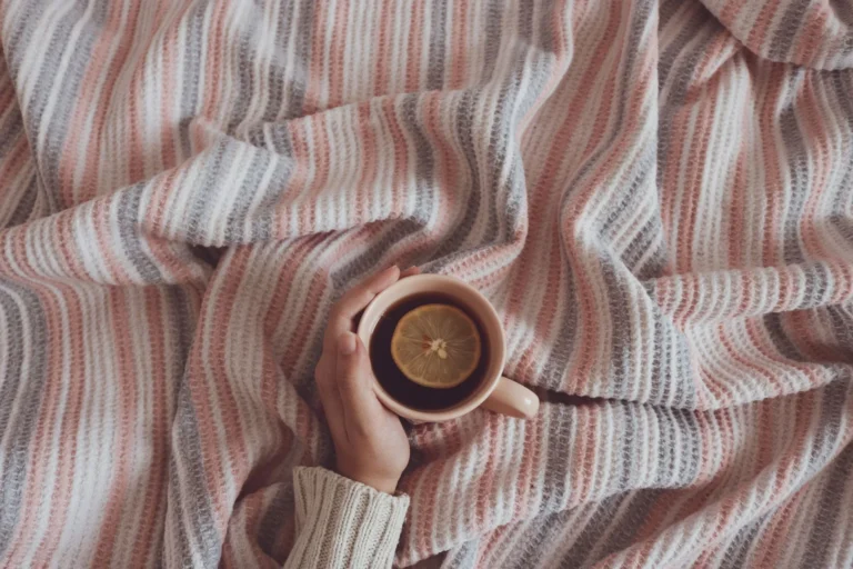A hand holding a cup of tea with a lemon slice on a striped blanket