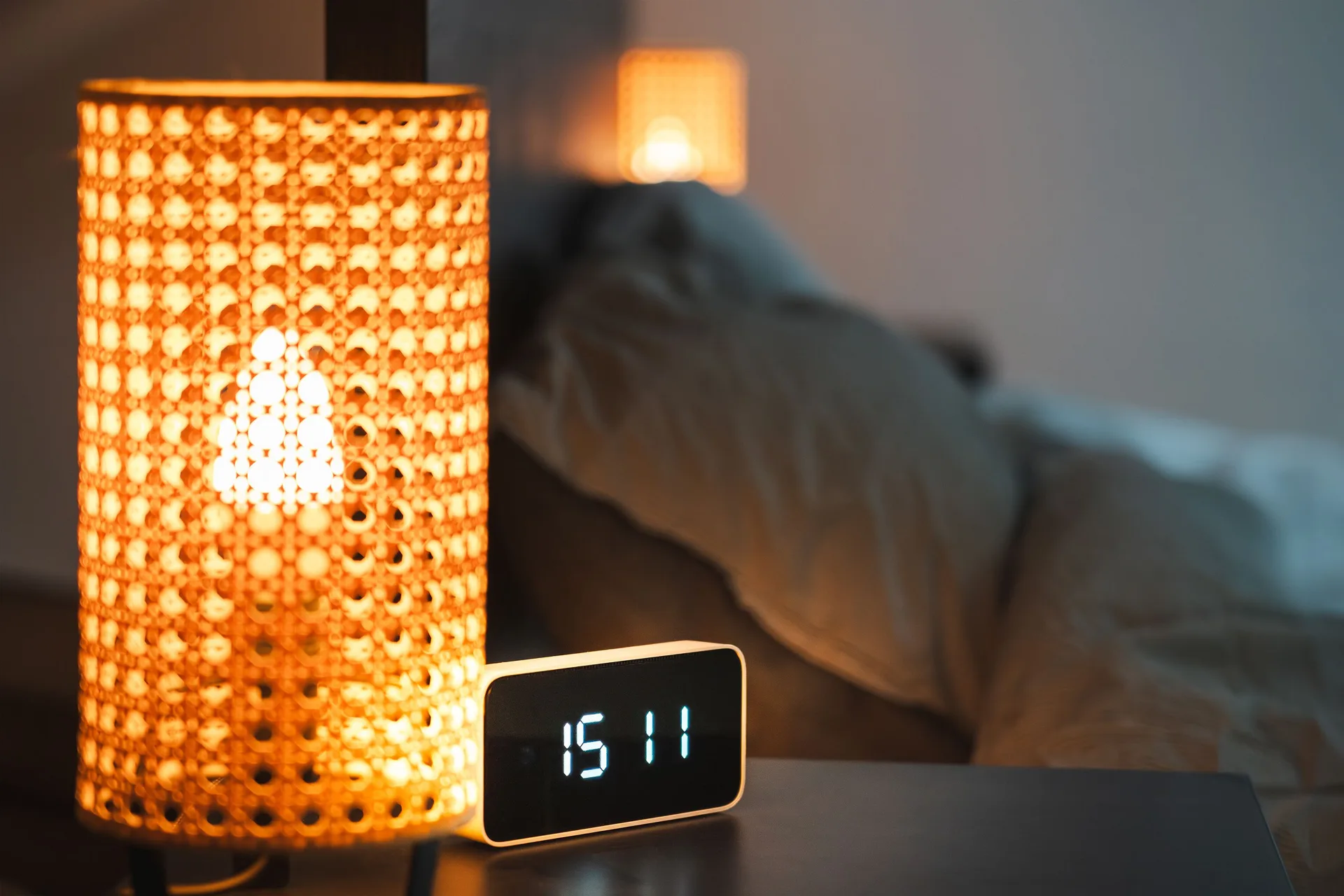 A glowing bedside lamp and a digital alarm clock on a nightstand beside a bed at night