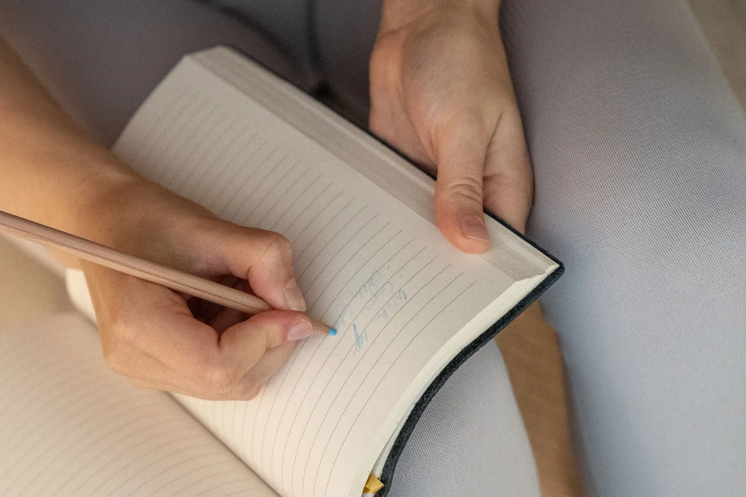 Hands writing with a pencil in an open lined notebook held on the lap