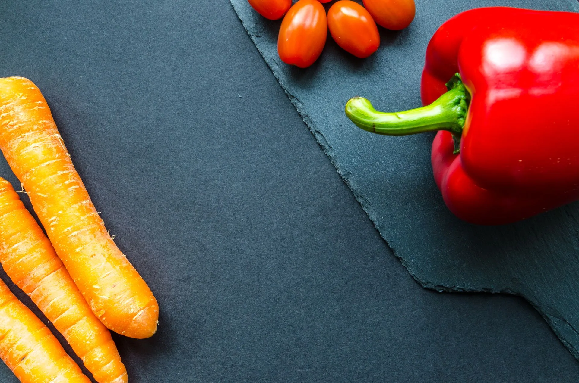 Fresh orange carrots, a red bell pepper and small tomatoes on a dark slate surface