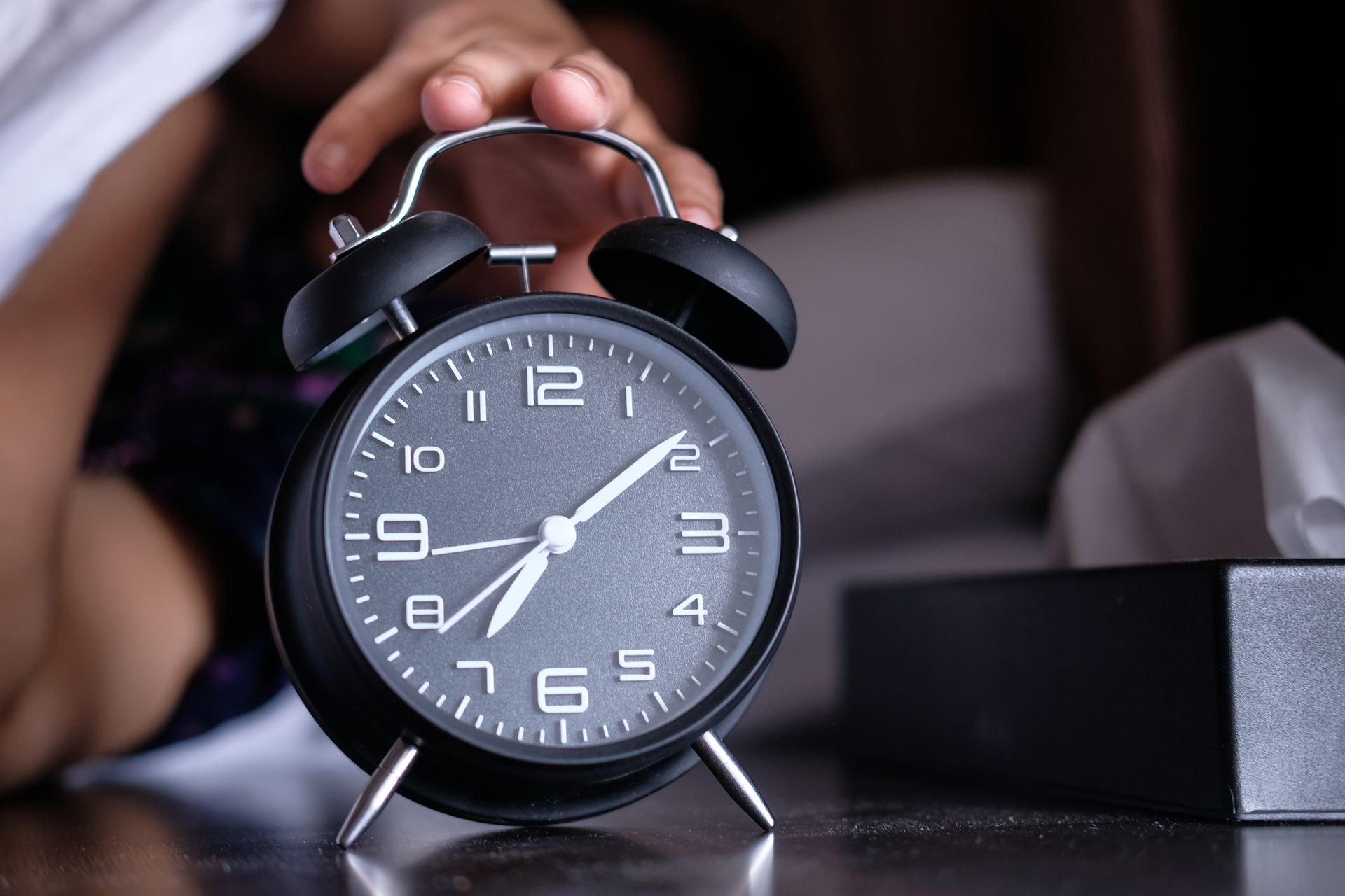 A hand reaching toward a black analog alarm clock on a bedside table