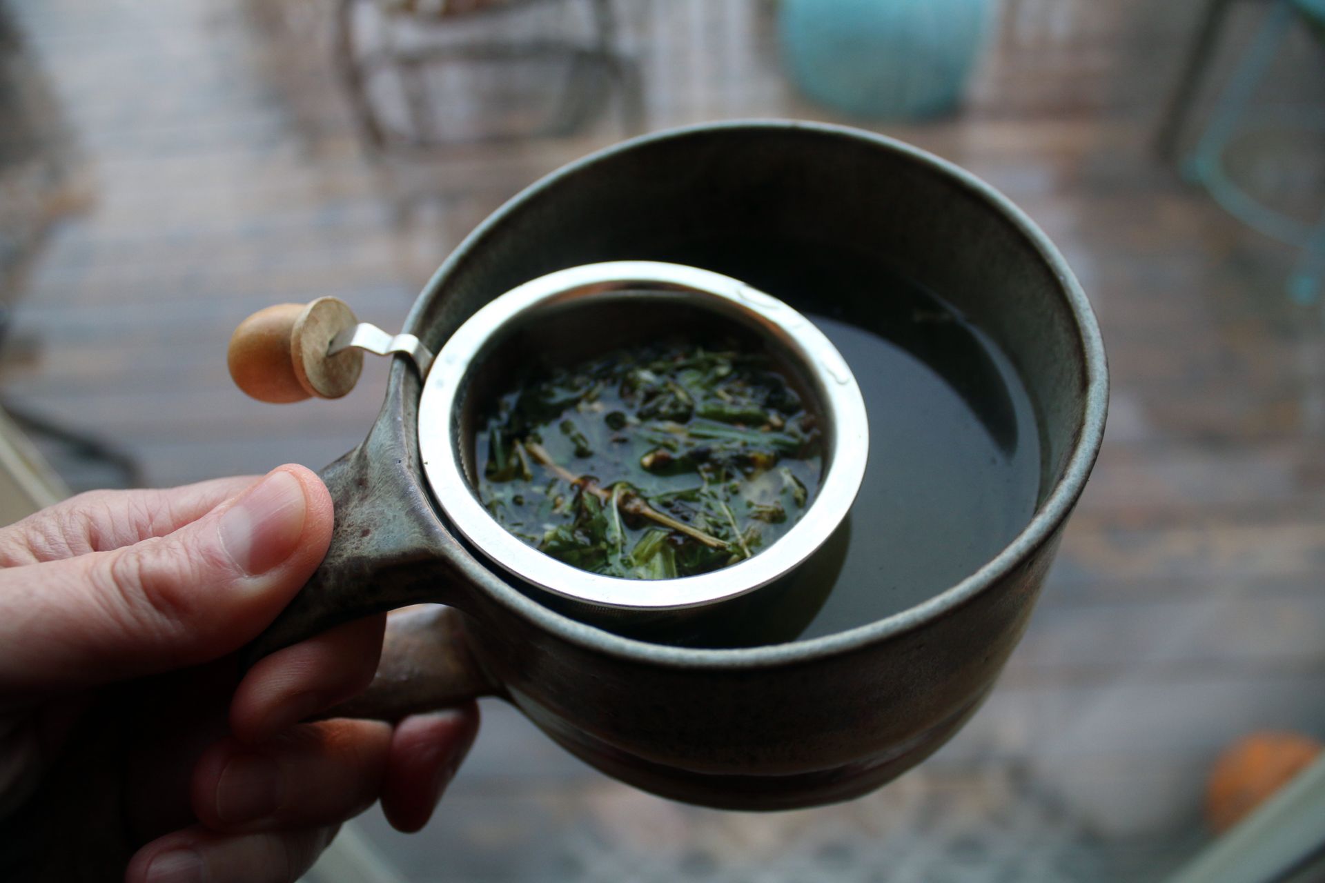 A hand holding a mug with a tea strainer full of loose herbs