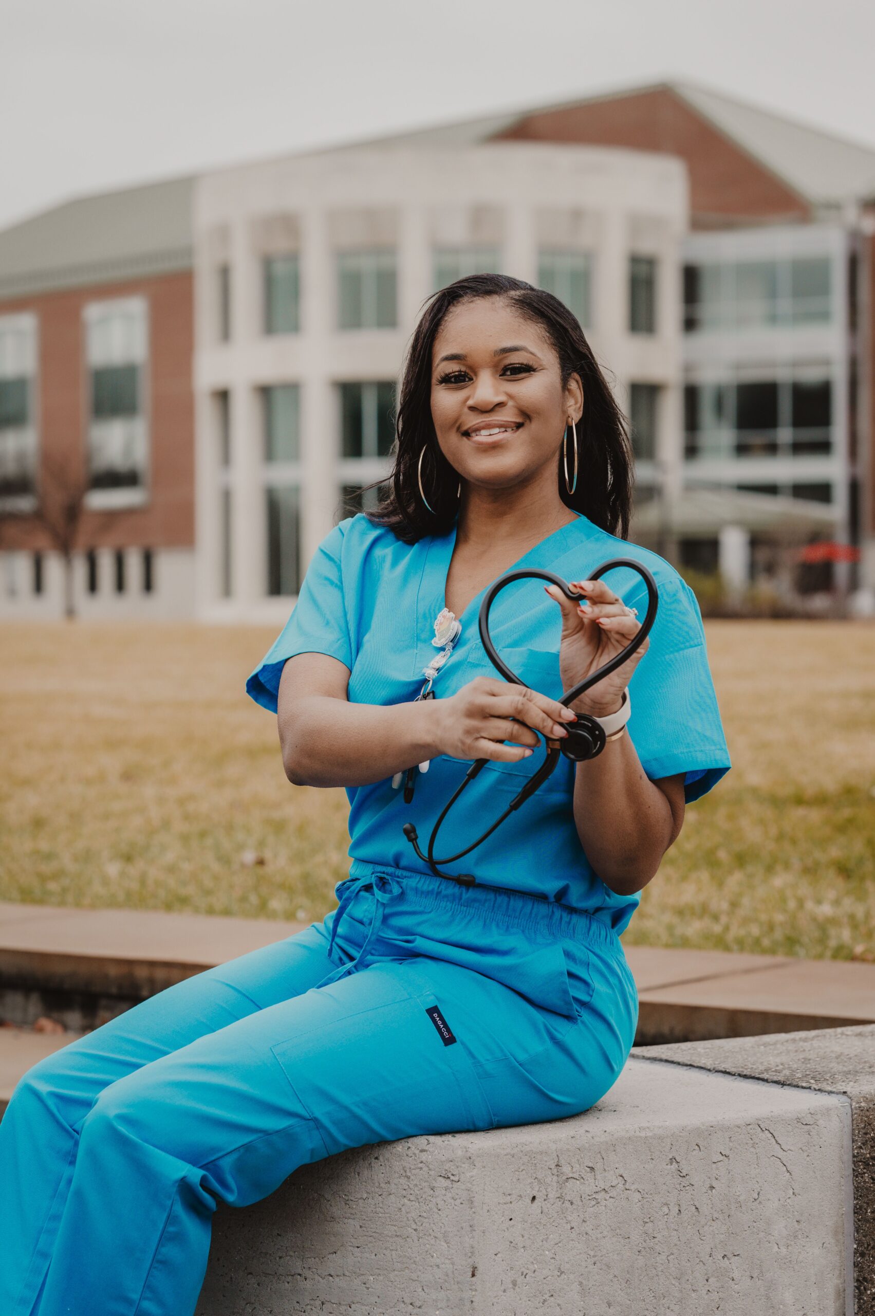 Smiling woman in blue scrubs forming a heart shape with a stethoscope outdoors