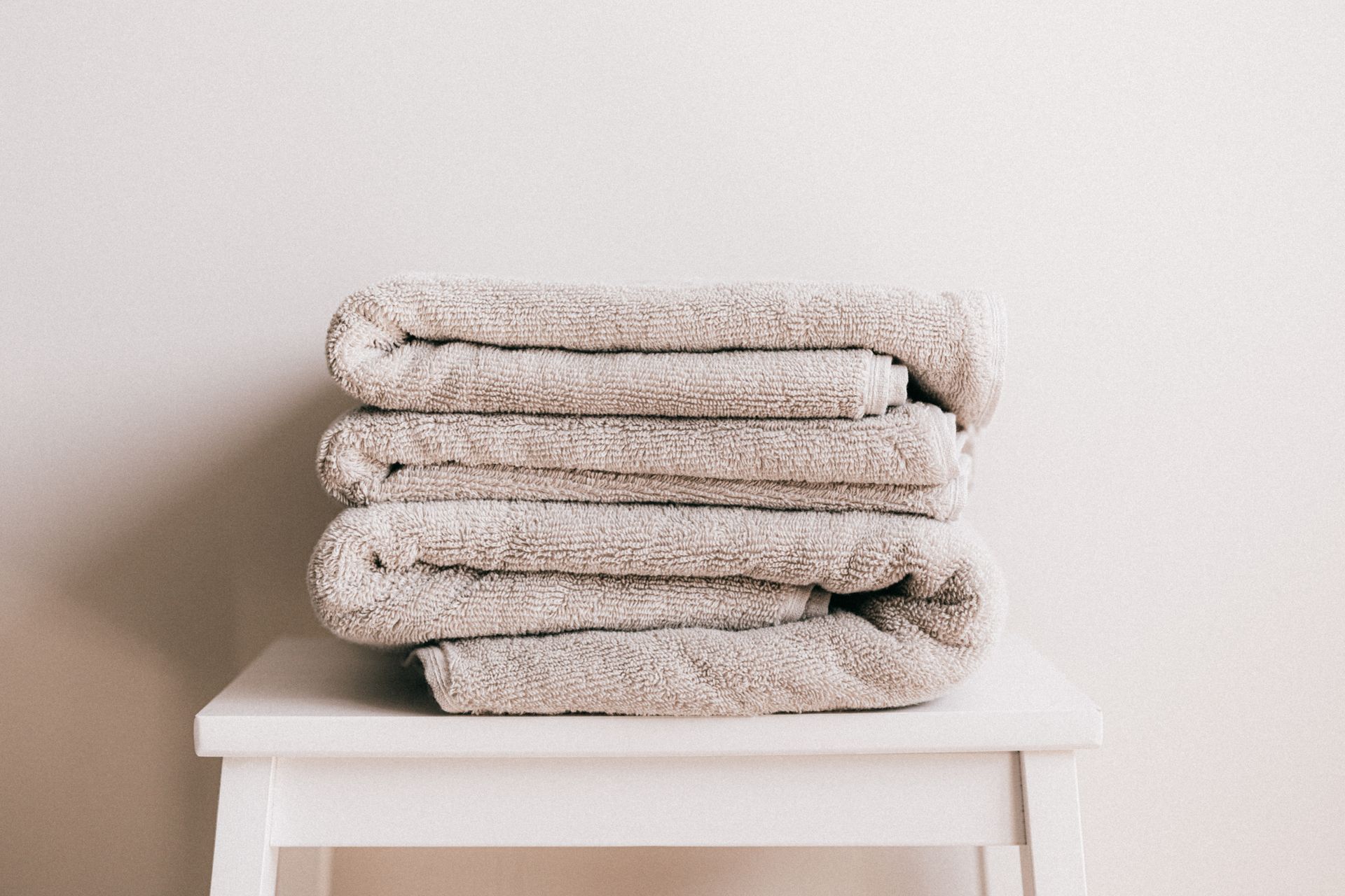 A neat stack of folded soft beige cotton towels on a small white stool against a plain pale wall