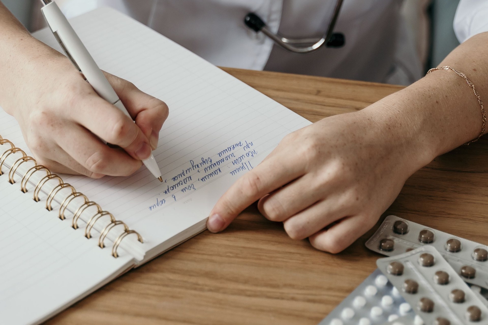 A doctor's hands writing notes in a notebook on a wooden desk with blister packs
