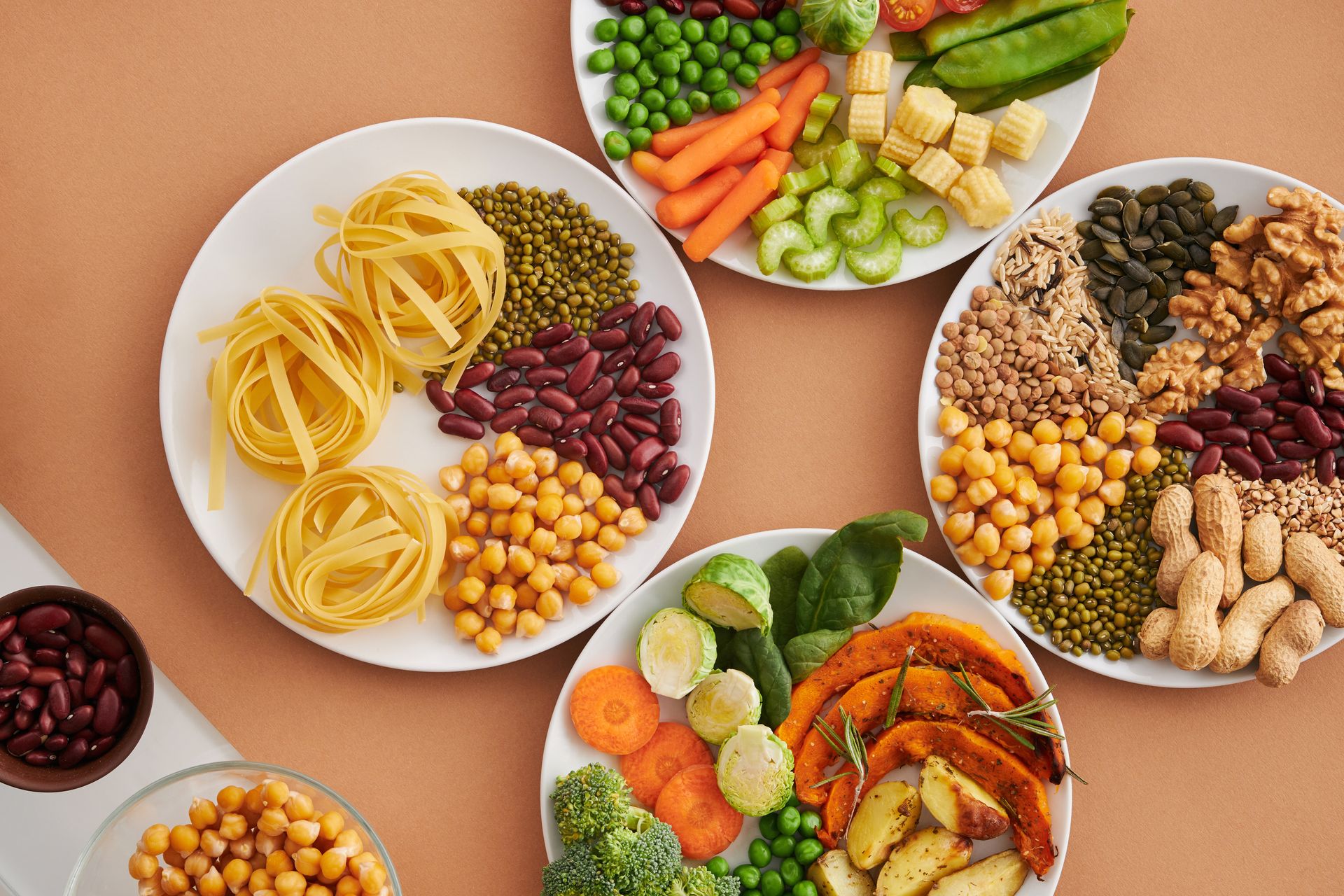 Overhead view of white plates holding chickpeas, lentils, kidney beans, green peas, spinach, Brussels sprouts, broccoli, roasted squash and nuts on a warm terracotta table