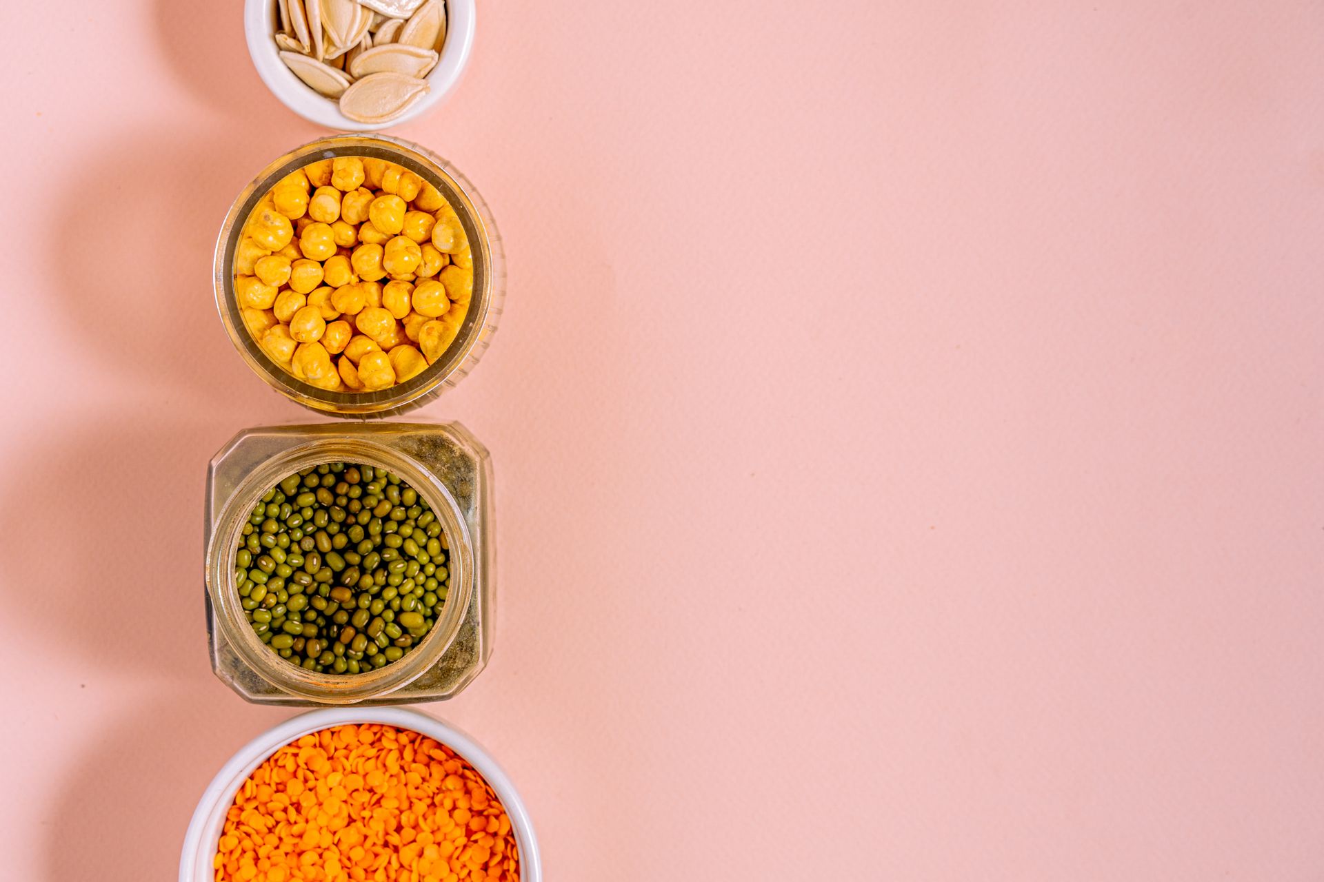 Overhead view of jars and bowls of chickpeas, mung beans, red lentils and pumpkin seeds on a pink background