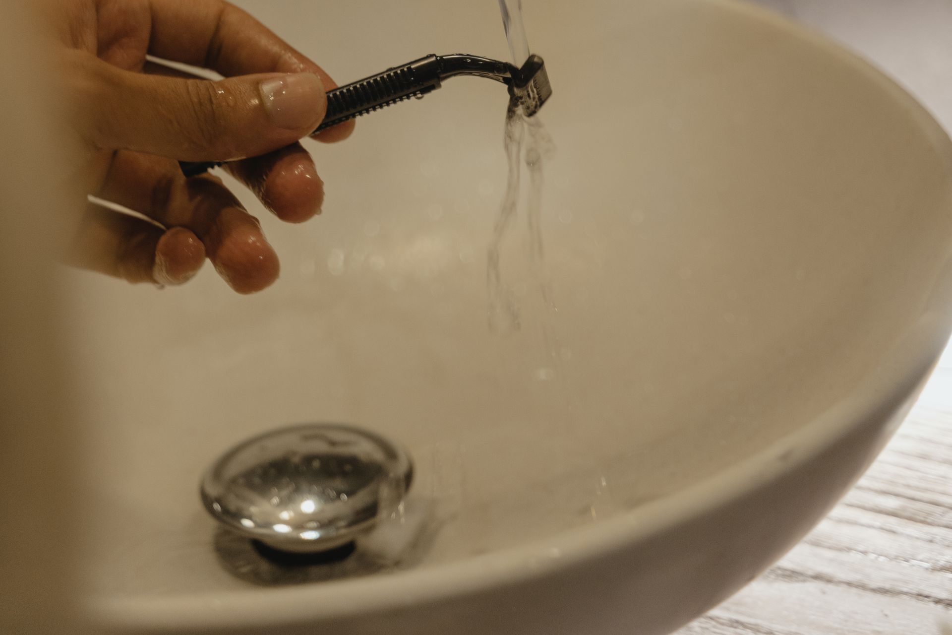 A hand rinsing a plain razor under running water at a bathroom sink.