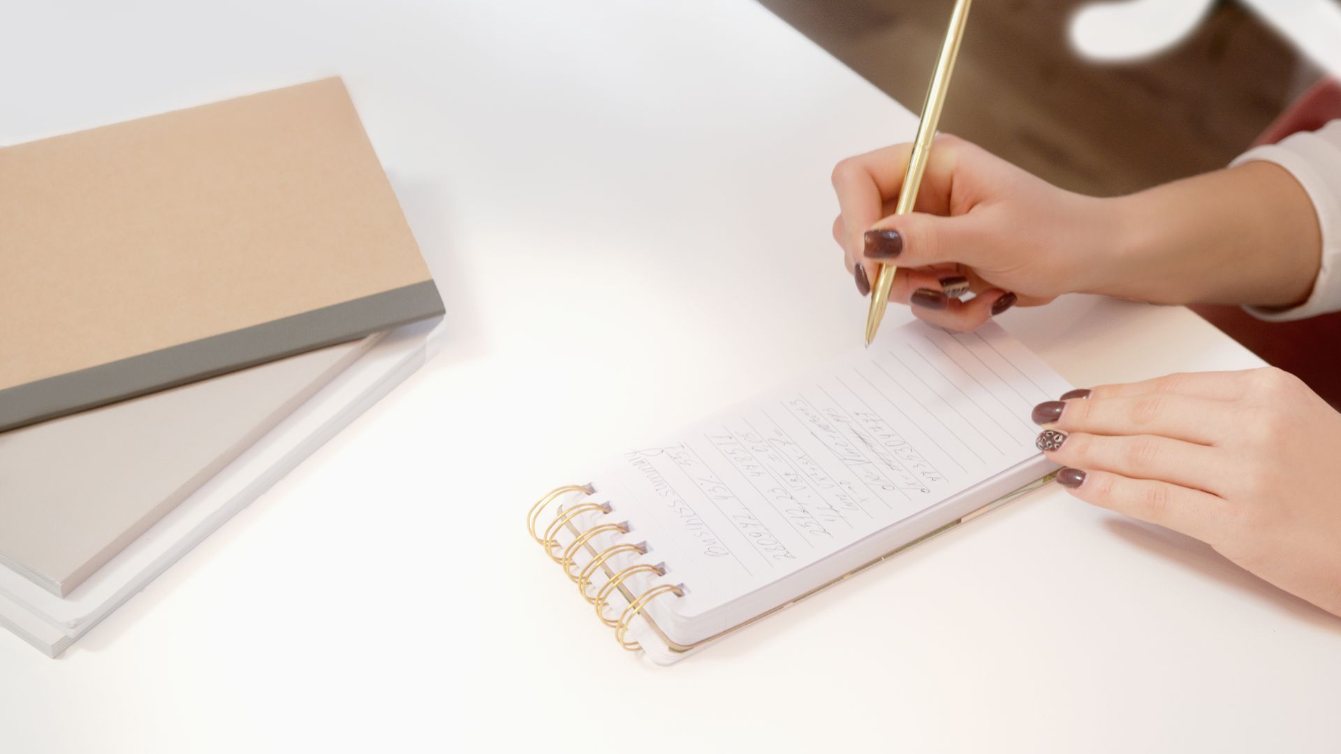 Hands writing notes in a small spiral notebook on a white desk