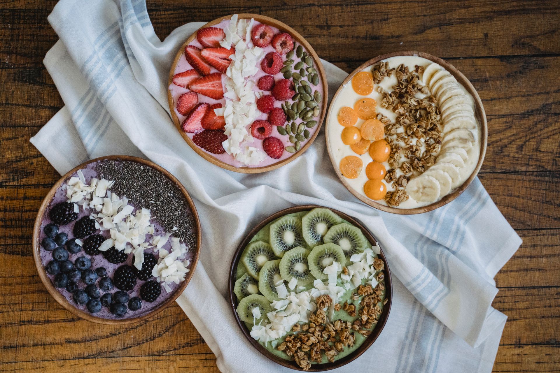 Four smoothie bowls topped with fruit and seeds on a wooden table