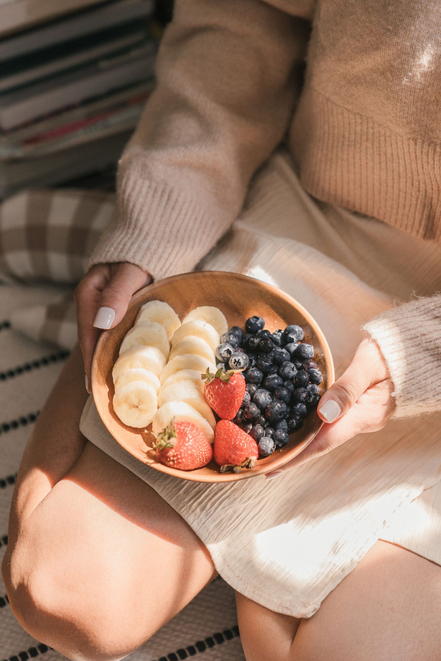 A wooden bowl of banana slices, strawberries and blueberries held in a lap