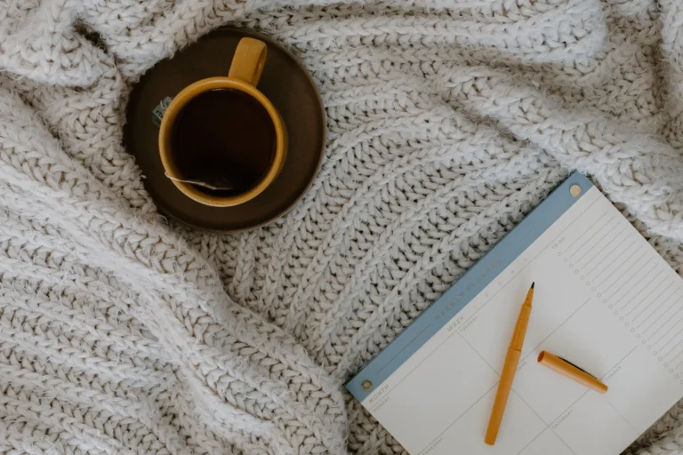 A cup of tea on a knitted blanket beside a weekly planner and pen