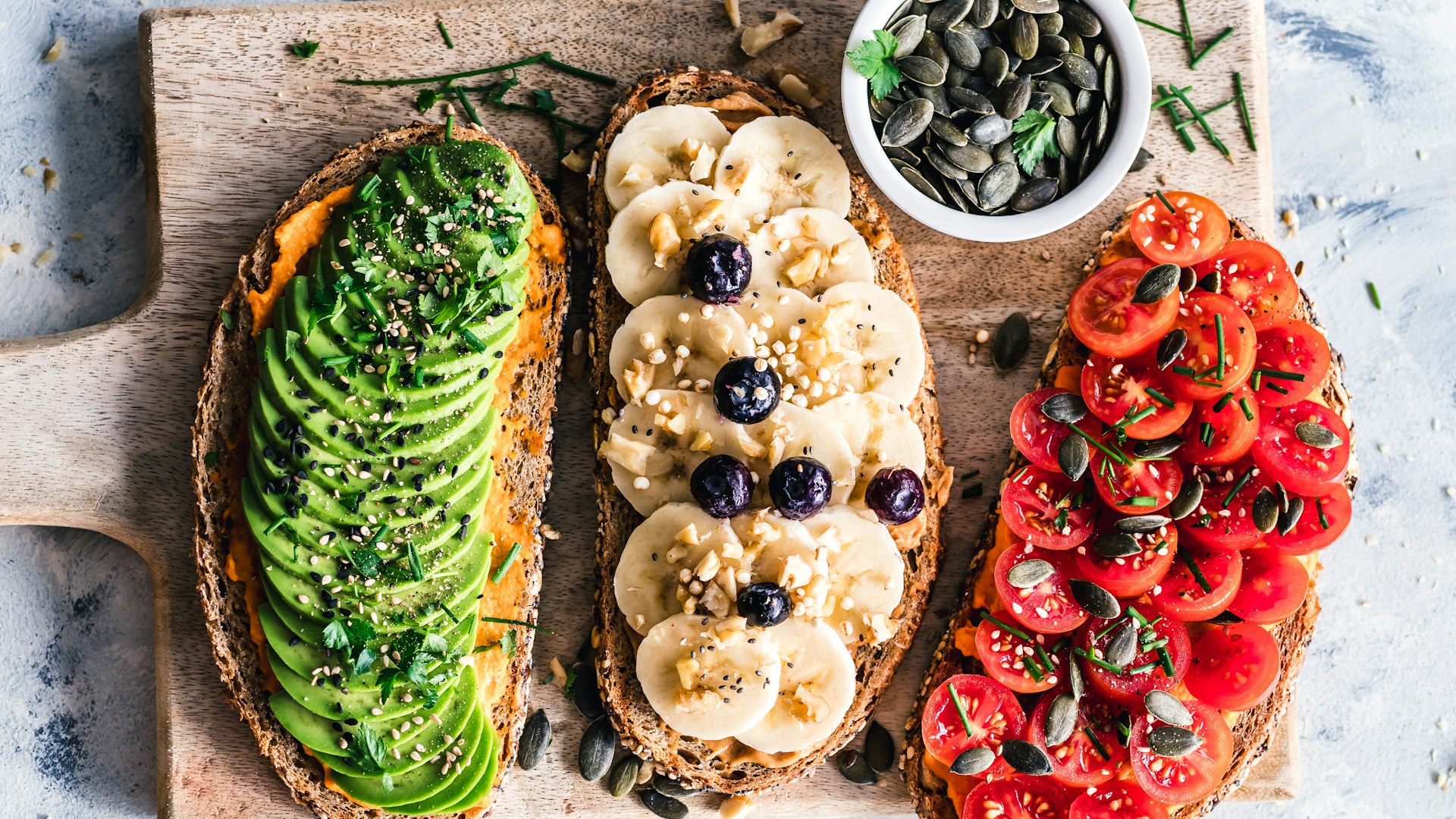Three slices of toast topped with avocado, banana with blueberries, and cherry tomatoes on a wooden board