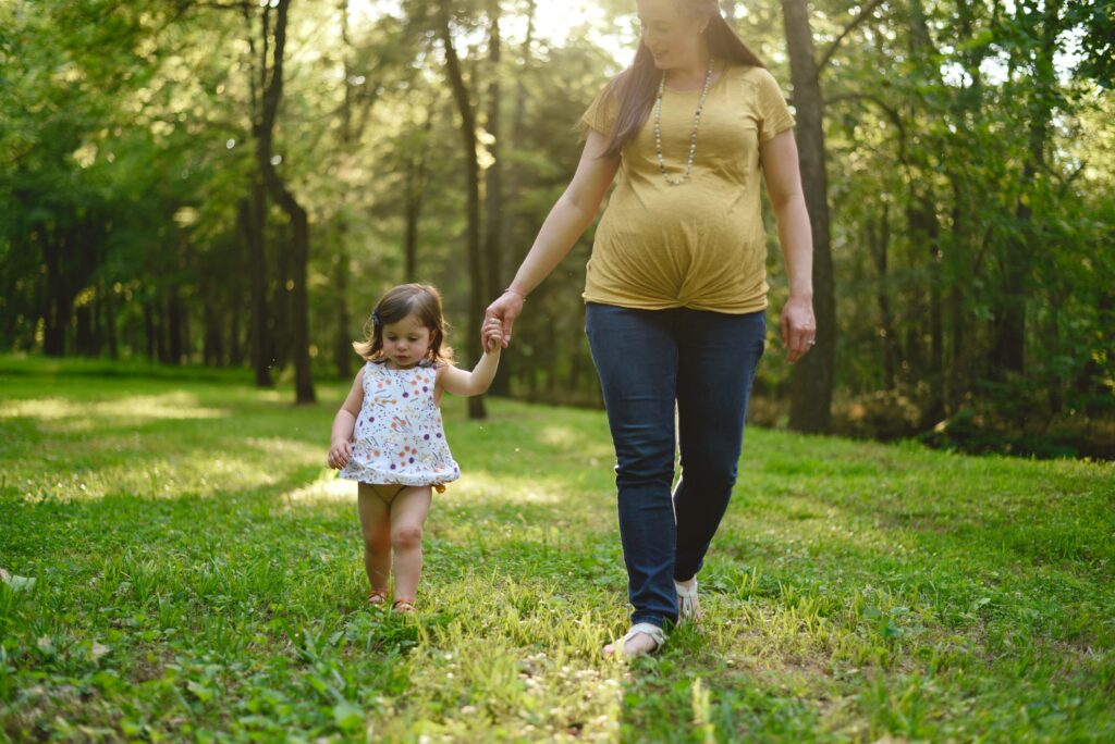 A pregnant woman walks hand-in-hand with her toddler daughter through a sunlit park.
