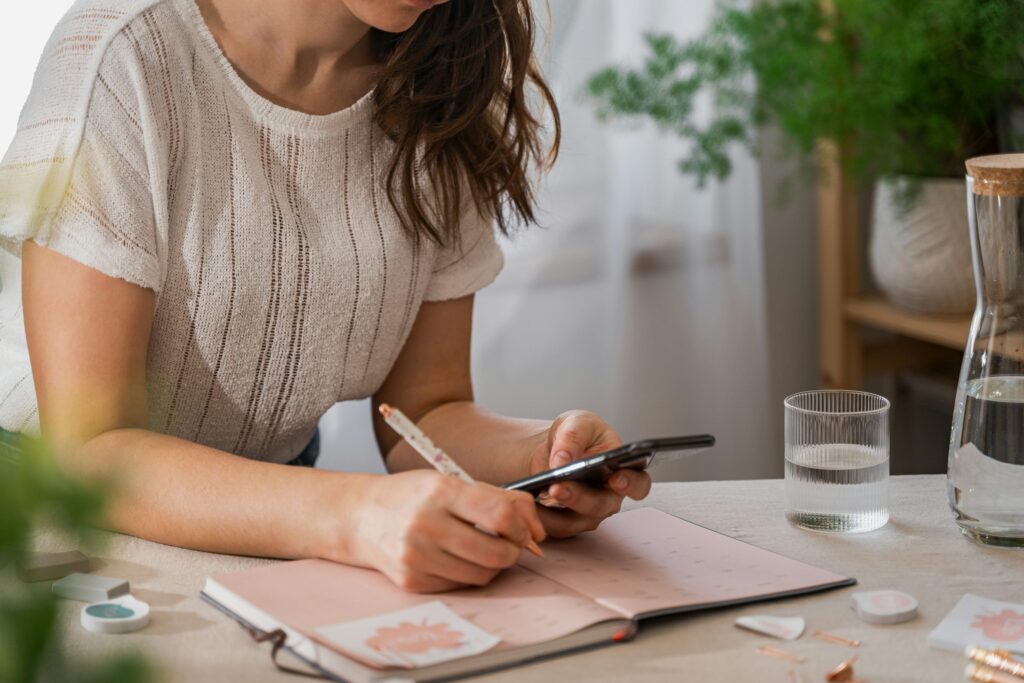 Woman writing in a dated planner while checking her phone, tracking her cycle at home