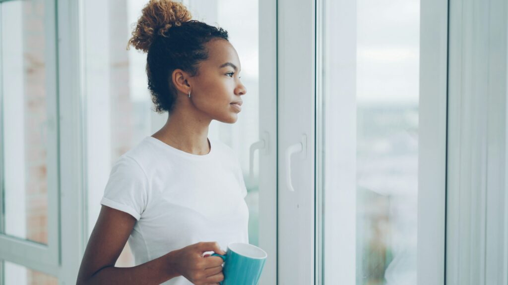 Woman holding a warm mug of coffee while looking out a sunlit window, considering whether coffee affects fertility