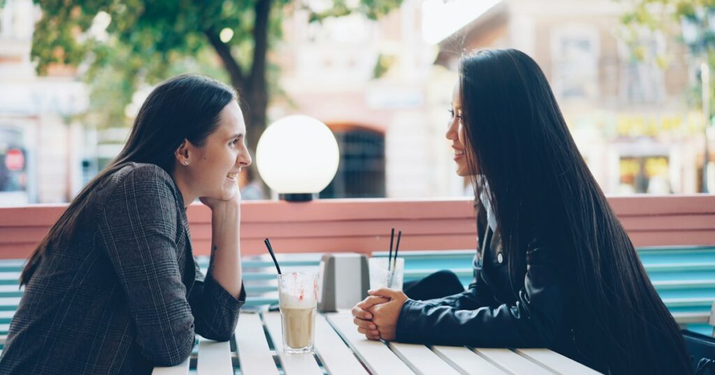 Two women sitting together at a café table, smiling and talking warmly during a relaxed conversation, illustrating an open conversation about herpes 1 vs herpes 2