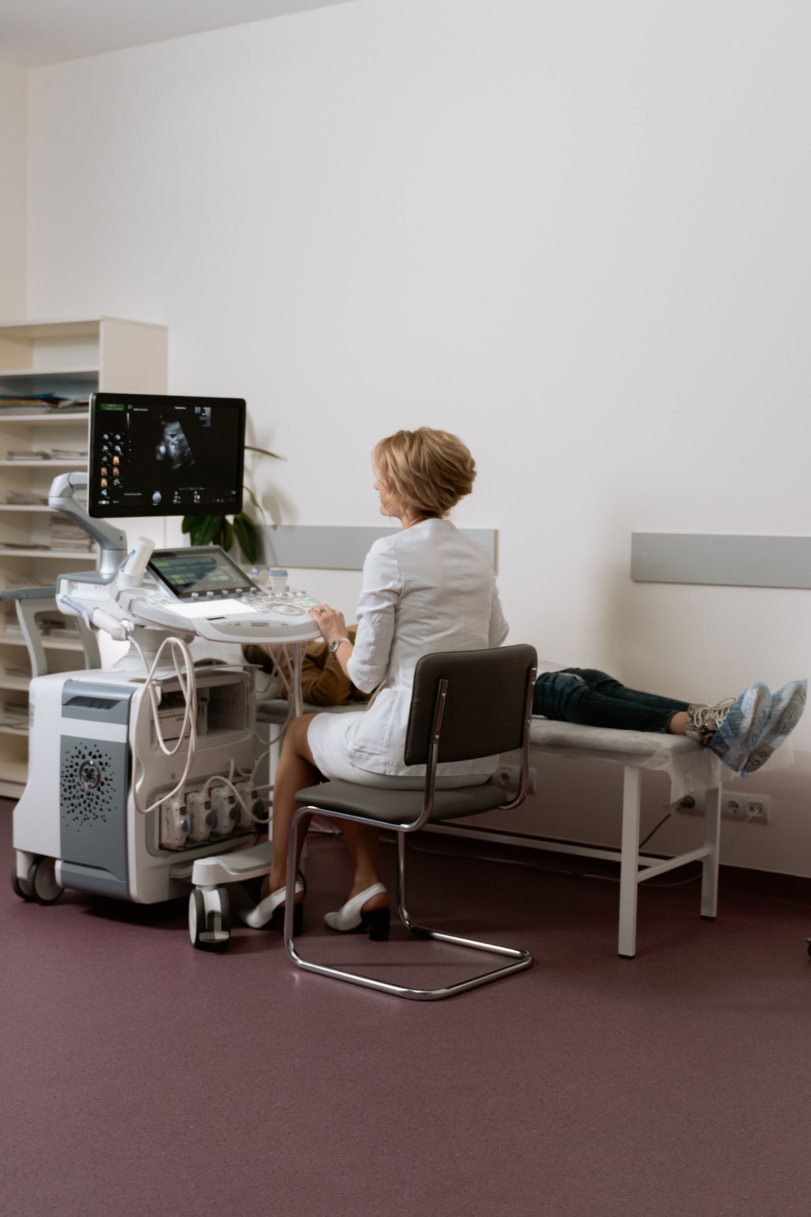 A sonographer seen from behind and a patient lying on an exam bed in a clinic room